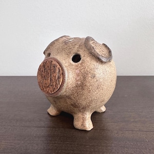 Peut inclure: Une tirelire en céramique tachetée de couleur beige. Le cochon a de petites oreilles arrondies, un museau circulaire à texture boisée et de petites pattes trapues. La tirelire a deux trous pour les yeux et une fente sur le dessus.