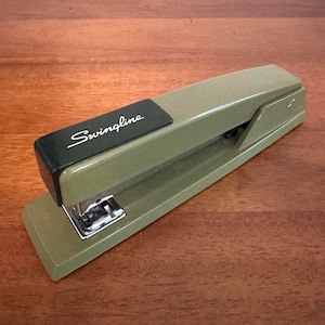 May include: A vintage olive green Swingline stapler with a black top plate featuring the white cursive brand name. The metal stapling mechanism is visible at the front, and the body has a smooth, matte finish with a long, rectangular shape.
