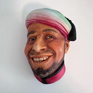 Peut inclure: Sculpture détaillée d'un visage d'homme souriant, portant un turban à rayures rouges, blanches et vertes. La sculpture a une barbe et est encadrée d'une bordure rouge et noire. Le visage a un teint réaliste.