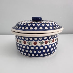 May include: A ceramic casserole dish with a lid. The dish is white with a blue, red, and green floral pattern. The lid has a dark blue knob. The dish is round and has a slightly flared rim.