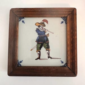 May include: A framed ceramic tile featuring a colorful illustration of a man playing a flute. The man is dressed in blue, black, green, and yellow attire, with a feathered hat and a sword. The tile is set in a dark wood frame with decorative corner accents.
