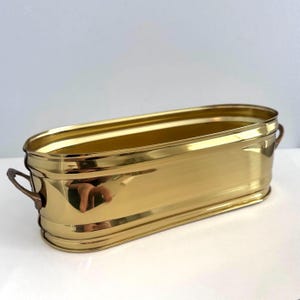 May include: A rectangular brass planter with rounded corners and two small handles. The planter has a polished, reflective surface, and is designed to hold plants or flowers. The overall design is simple and elegant.