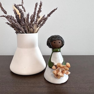 May include: A white ceramic vase contains dried lavender. A small ceramic doll with a black bob, green collar, and white dress holds dried flowers. The items are on a dark brown surface.