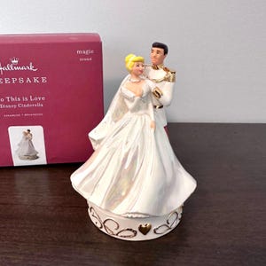 May include: A Hallmark Keepsake ornament depicting Cinderella and Prince Charming in wedding attire. The figurine is white with iridescent highlights, and the base has gold detailing. The box reads "So This is Love Disney Cinderella".
