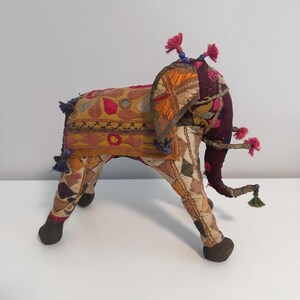 May include: A decorative, stuffed elephant figurine with intricate embroidery. The elephant features a patchwork design in shades of yellow, orange, and burgundy, with pink and blue accents. It has a long trunk and is adorned with tassels.