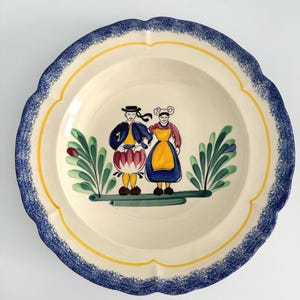 May include: A vintage ceramic soup bowl with a blue and yellow rim. The bowl features a hand-painted design of a couple in traditional clothing standing in front of green foliage.
