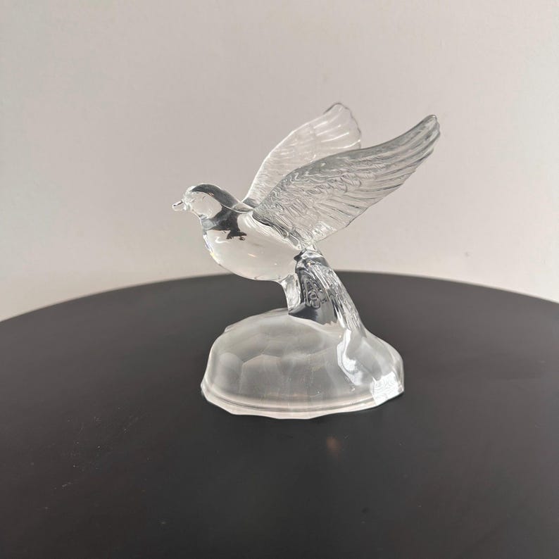 Puede incluir: Una figurita de cristal transparente de un p&aacute;jaro con las alas extendidas, posada sobre una peque&ntilde;a base. El p&aacute;jaro est&aacute; en vuelo, con la cabeza girada hacia un lado.