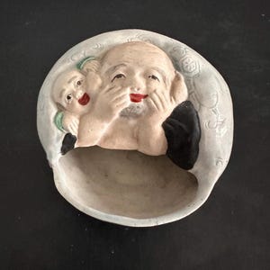 Peut inclure: Un plat en céramique blanche avec un motif en relief de deux personnages souriants. Le personnage le plus grand a une barbe et se couvre la bouche avec ses mains. Le personnage le plus petit est caché derrière le personnage le plus grand. Le plat est recouvert d'un glaçage bleu clair et d'un motif floral.