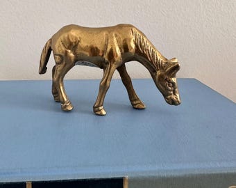 May include: A small, vintage brass horse figurine. The horse is in a grazing position, with its head lowered. The figurine has a detailed mane and tail, and is displayed on a blue surface.