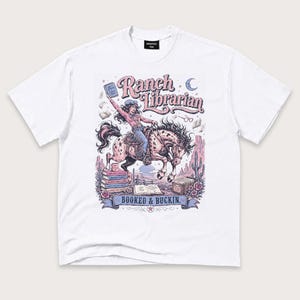 Puede incluir: Una camiseta blanca de manga corta con un estampado gráfico de una persona montando un caballo moteado. El texto dice Ranch Librarian y Booked & Buckin. La ilustración incluye pilas de libros, una luna creciente, gafas y cactus.
