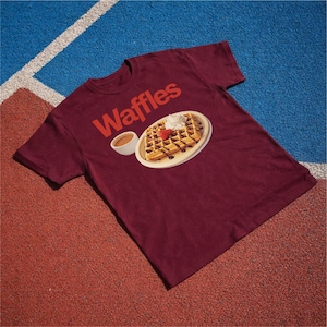 以下が含まれることがあります： 赤色の「Waffles」の文字が入った、マルーン色のTシャツ。ホイップクリーム、イチゴ、チョコレートシロップを添えたワッフルのグラフィックが描かれており、お皿と小さなシロップのカップが描かれています。