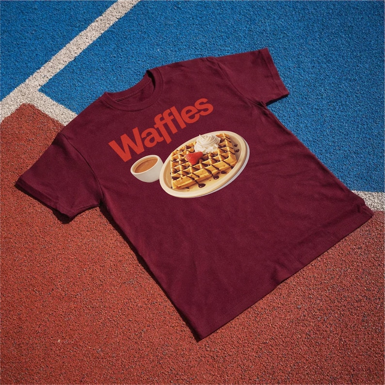 以下が含まれることがあります： 赤色の「Waffles」の文字が入った、マルーン色のTシャツ。ホイップクリーム、イチゴ、チョコレートシロップを添えたワッフルのグラフィックが描かれており、お皿と小さなシロップのカップが描かれています。