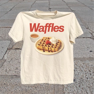 T-shirt featuring bold red Waffles text above a plated waffle with whipped cream, chocolate drizzle, strawberry, and coffee.