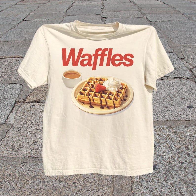 T-shirt featuring bold red Waffles text above a plated waffle with whipped cream, chocolate drizzle, strawberry, and coffee.