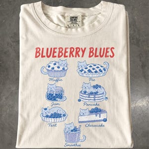 May include: A cream-colored t-shirt featuring the red text BLUEBERRY BLUES and blue illustrations of cats integrated with blueberry-themed foods labeled Muffin, Pie, Jam, Pancake, Tart, Cheesecake, and Smoothie, with a Comfort Colors neck label.