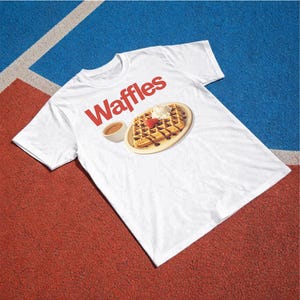以下が含まれることがあります： 赤い文字で「Waffles」と書かれた白いTシャツ。チョコレートシロップ、ホイップクリーム、イチゴが乗ったワッフルのグラフィックと、小さなシロップのボウルが描かれています。