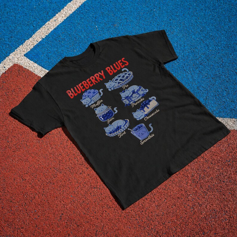 May include: A black short-sleeve t-shirt featuring the red text BLUEBERRY BLUES and blue illustrations of cats shaped like food items. The graphic includes labels for Muffin, Pie, Pancake, Jam, Cheesecake, Tart, and Smoothie, each depicting a cat.
