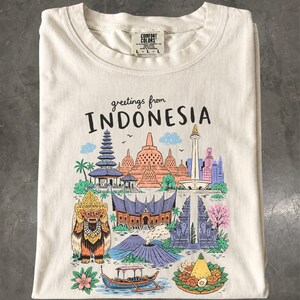 Könnte beinhalten: Ein cremefarbenes T-Shirt mit einem bunten Grafikdruck und dem Text "greetings from INDONESIA". Die Illustration zeigt verschiedene Wahrzeichen, eine traditionelle Maske, einen Vulkan, ein Boot und ein Gericht in Blau-, Rosa- und Orangetönen.