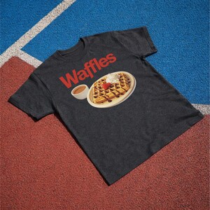 以下が含まれることがあります： 赤色の「Waffles」の文字が入った濃いグレーのTシャツ。チョコレートシロップ、ホイップクリーム、イチゴが乗ったワッフルのグラフィックと、小さなシロップのカップが描かれています。