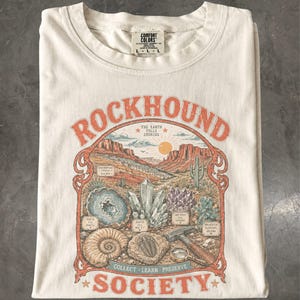 以下が含まれることがあります： 「ROCKHOUND SOCIETY」および「THE EARTH TELLS STORIES」という文字がプリントされたクリーム色のTシャツ。イラストには砂漠の風景、様々な結晶、化石、地質調査用ハンマー、拡大鏡が描かれています。