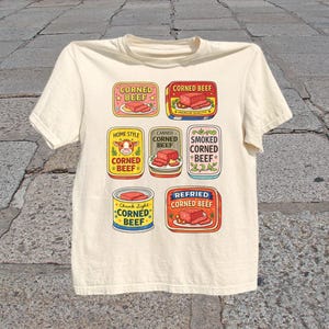 May include: Cream-colored t-shirt featuring colorful illustrations of corned beef labels. The labels include text such as "Corned Beef," "Home Style," and "Refried Corned Beef." The design is a playful, vintage-inspired graphic.