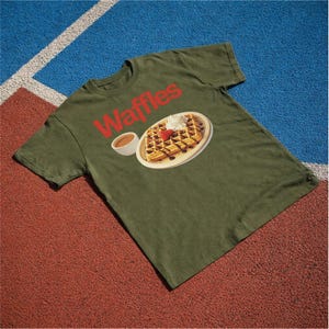 以下が含まれることがあります： 赤色の「Waffles」の文字が入ったオリーブグリーンのTシャツ。ホイップクリーム、イチゴ、チョコレートシロップを添えたワッフルのグラフィックが描かれており、お皿とシロップのカップが描かれています。