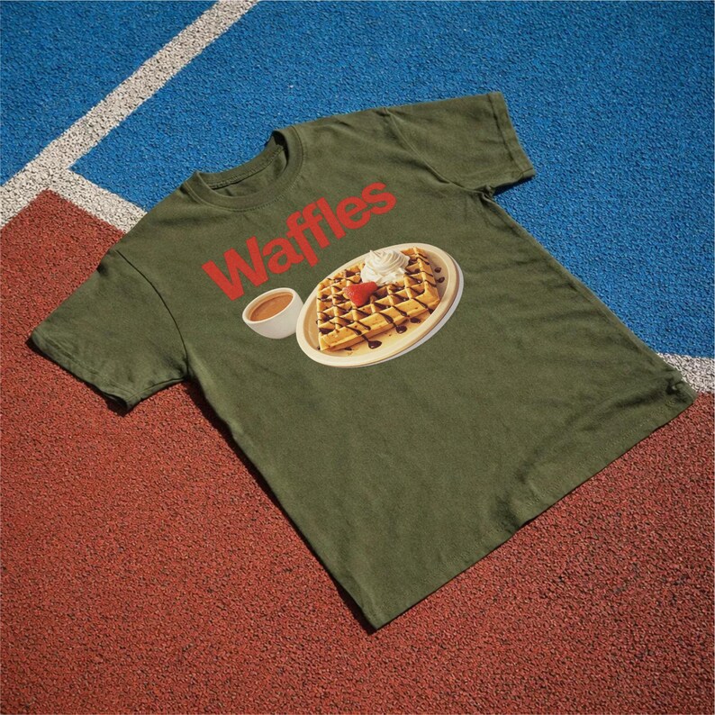 以下が含まれることがあります： 赤色の「Waffles」の文字が入ったオリーブグリーンのTシャツ。ホイップクリーム、イチゴ、チョコレートシロップを添えたワッフルのグラフィックが描かれており、お皿とシロップのカップが描かれています。