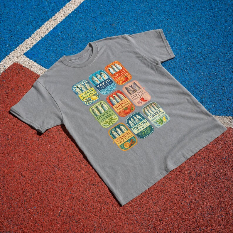 May include: A grey short-sleeve t-shirt featuring a graphic print of ten colorful rectangular sardine tin labels. Each label displays fish illustrations and text including Lisbon, Tokyo, Morocco, Basque, Napoli, Provence, Portland, and Istanbul.