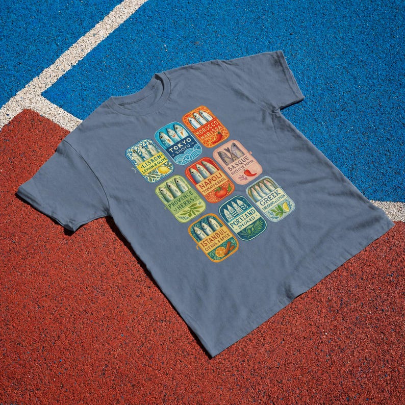 May include: A slate blue short-sleeve t-shirt featuring a graphic print of nine rectangular sardine tin labels. Each label displays a city name, including Tokyo, Lisbon, Morocco, Basque, Napoli, Provence, Portland, Greek, and Istanbul, with fish illustrations.