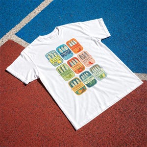 May include: A white short-sleeve t-shirt featuring a graphic print of nine colorful rectangular sardine tin labels. Each label displays a city name, flavor profile, and illustrations of fish, including Lisbon, Tokyo, Morocco, Basque, Napoli, Provence, Portland, Greek, and Istanbul.