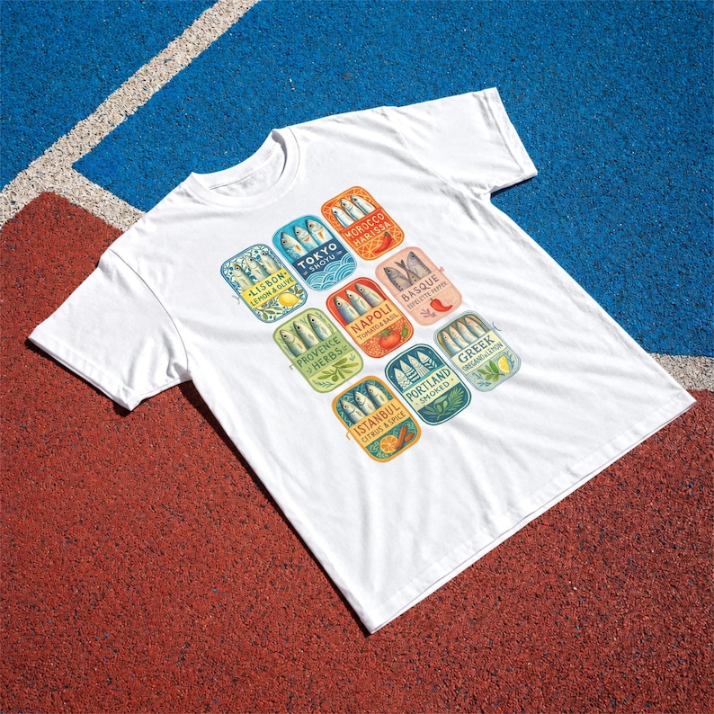 May include: A white short-sleeve t-shirt featuring a graphic print of nine colorful rectangular sardine tin labels. Each label displays a city name, flavor profile, and illustrations of fish, including Lisbon, Tokyo, Morocco, Basque, Napoli, Provence, Portland, Greek, and Istanbul.