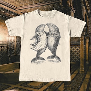 May include: Cream-colored t-shirt featuring a whimsical illustration of two whales in formal attire, embracing. One whale wears a dress, the other a bow tie. The graphic is in black and white, creating a vintage aesthetic.