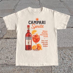 Könnte beinhalten: Cremefarbenes T-Shirt mit einem Campari Spritz-Design. Die Grafik zeigt eine Campari-Flasche, ein Cocktailglas mit Eis und einer Orangenscheibe sowie den Text "Campari Spritz". Zusätzlicher Text lautet "Bitter, Bubbly, Gloriously Fresh".