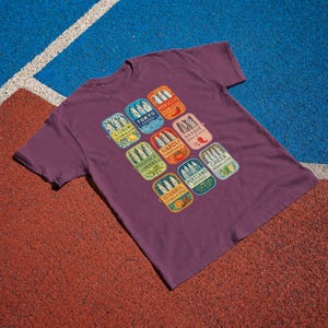 May include: A dark purple short-sleeve t-shirt featuring a graphic print of nine rectangular sardine tin labels. Each label displays a city name, including Lisbon, Tokyo, Morocco, Basque, Napoli, Provence, Portland, Greek, and Istanbul, with fish illustrations.