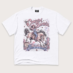 Könnte beinhalten: Ein weißes Kurzarm-T-Shirt mit einer grafischen Illustration einer Cowgirl auf einem Pferd, die eine Kaffeetasse hält. Der Text lautet Cowgirl Barista und Giddy Up & Grind. Das Design zeigt Kaffeebohnen, eine Kaffeekanne und eine Wüstenlandschaft.