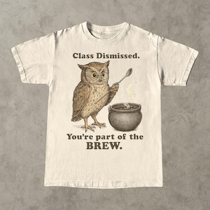 Peut inclure: T-shirt crème avec une illustration détaillée d'un hibou portant des lunettes et tenant une cuillère au-dessus d'un chaudron bouillonnant. Le texte dit "Class Dismissed. You're part of the BREW."