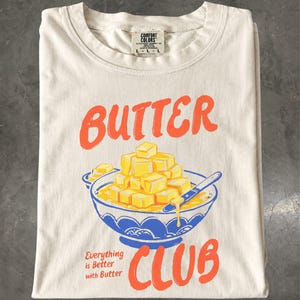 May include: A cream-coloured t-shirt featuring a graphic of a blue bowl filled with yellow butter cubes and a butter knife. The text reads "BUTTER CLUB" in orange and "Everything is Better with Butter" in smaller orange font. The shirt has a Comfort Colors label.