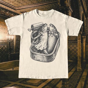 Könnte beinhalten: Cremefarbenes T-Shirt mit einer Schwarz-Weiß-Illustration von zwei anthropomorphen Fischen in einer Sardinenbüchse. Einer der Fische trägt ein Kleid, der andere eine Fliege. Die Dose hat eine Lasche.