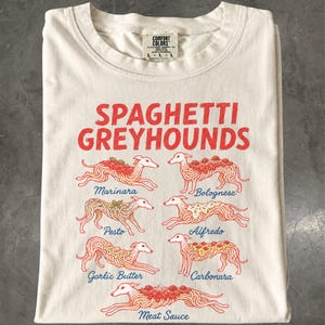 May include: A cream-colored t-shirt featuring the red text SPAGHETTI GREYHOUNDS and seven illustrations of greyhounds with various pasta dishes on their backs, labeled Marinara, Bolognese, Pesto, Alfredo, Garlic Butter, Carbonara, and Meat Sauce.