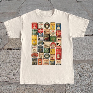 May include: Cream-colored t-shirt featuring a collage of vintage coffee tin labels. The labels are in various colors and designs, showcasing brands like "Caffe Lusso," "Folgers," and "Moka." The shirt has a classic crew neck and short sleeves.