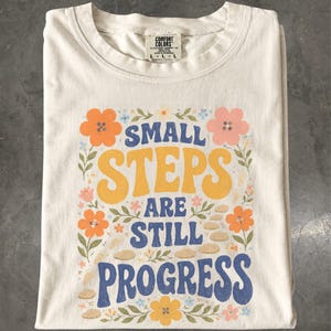 Könnte beinhalten: Ein cremefarbenes T-Shirt mit einem Grafikdruck, der den Text "SMALL STEPS ARE STILL PROGRESS" in Blau und Gelb zeigt. Das Design ist umgeben von orangefarbenen, rosa und gelben Blumen, grünen Blättern und kleinen beigen Steinen auf hellem Grund.