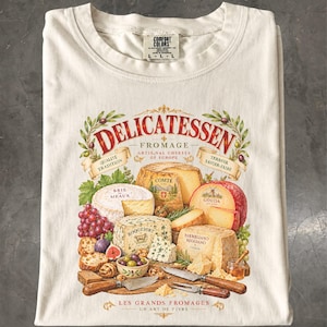 May include: An off-white t-shirt featuring a graphic print of various cheeses, including Brie de Meaux, Comte, Roquefort, Gouda, and Parmigiano Reggiano, with the text Delicatessen, Les Grands Fromages, and other culinary labels.