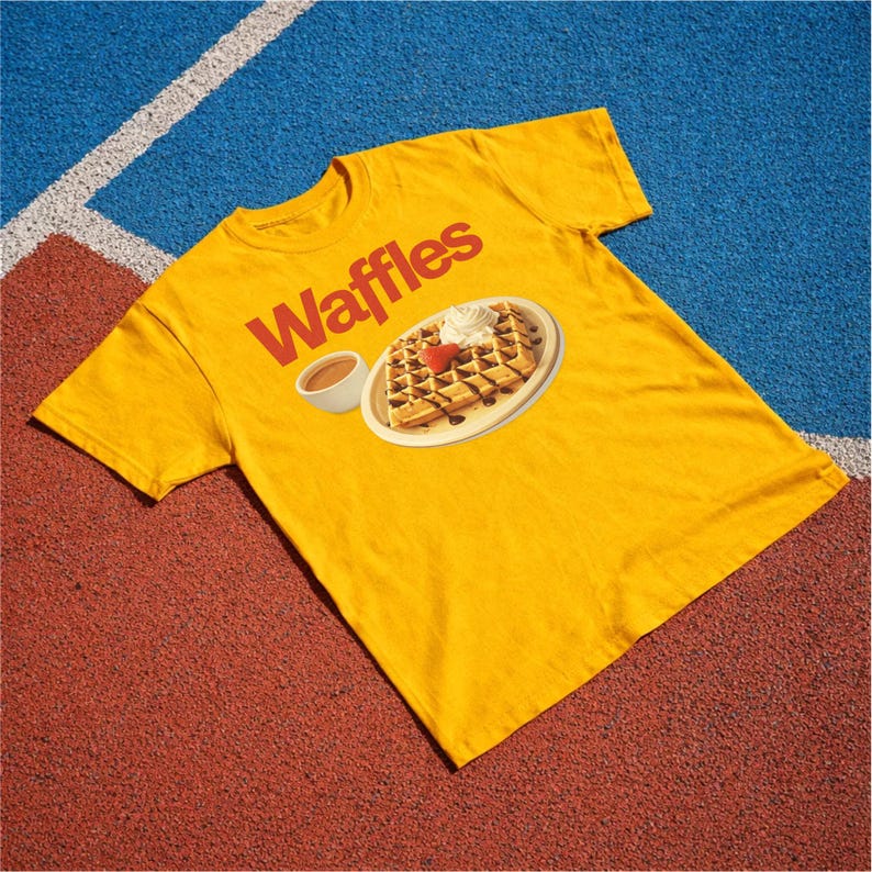 以下が含まれることがあります： 赤色のブロック文字で「Waffles」と書かれたマスタードイエローのTシャツ。ホイップクリーム、イチゴ、チョコレートシロップを添えたワッフルのグラフィックが描かれており、お皿と小さなシロップのカップが描かれています。