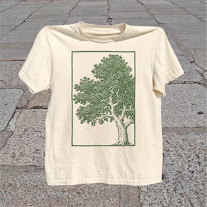Könnte beinhalten: Cremefarbenes T-Shirt mit einer grünen Baumillustration in einem rechteckigen Rahmen. Der Baum hat ein detailliertes Design mit strukturiertem Stamm und belaubten Ästen. Das Shirt besteht aus weichem Material und hat kurze Ärmel.