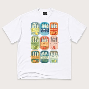 May include: A white short-sleeve t-shirt featuring a graphic print of nine rectangular sardine tin illustrations. Each tin displays a different city name, flavor profile, and fish arrangement, including Lisbon, Tokyo, Morocco, Provence, Napoli, Basque, Istanbul, Portland, and Greek.