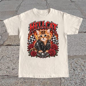 May include: Off-white t-shirt with a graphic of a kitten in a leather jacket, roses, flames, and checkered flags. The word "HELLCAT" is in a fiery font. The design includes skulls. The t-shirt is a casual wear item.
