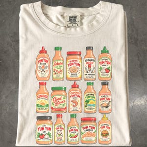 Puede incluir: Una camiseta de color crema con un estampado gráfico de quince botellas de salsa diferentes dispuestas en tres filas. Cada botella tiene una etiqueta única con texto como Yum Yum Sauce, Sakura, Hibachi Magic, Sunrise y Lucky Yum Yum, junto a ilustraciones.