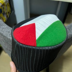 Puede incluir: Un sombrero negro de tela con estampado y una parte superior de tela roja, blanca y verde. La parte superior es una bandera palestina.