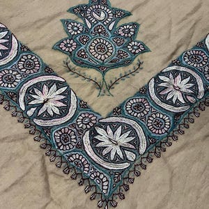 Puede incluir: Un chal beige con bordados intrincados en tonos azules, rosas y blancos. El bordado presenta motivos florales y paisley.
