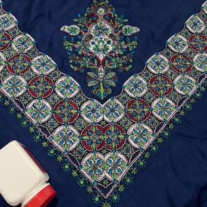 Puede incluir: Primer plano de un textil azul marino con intrincados patrones bordados en rojo, verde, blanco y azul. El diseño presenta motivos florales y geométricos. Un recipiente blanco con tapa roja está en la esquina inferior izquierda.